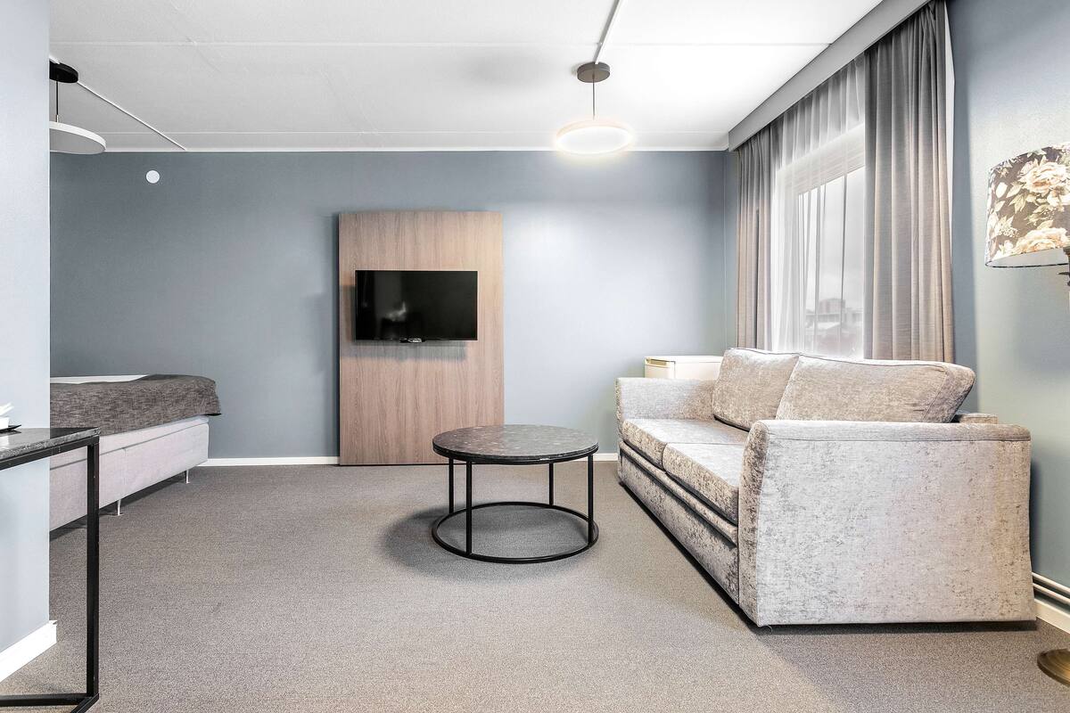 A modern living room with a round coffee table, a sofa, a TV mounted on a wooden panel, and a large window with curtains.