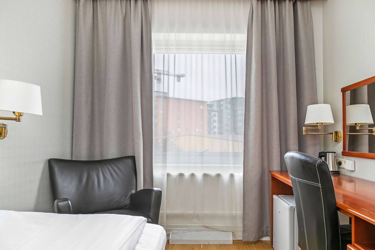A hotel room with a bed, a desk with a chair, a radiator, and a window with curtains.