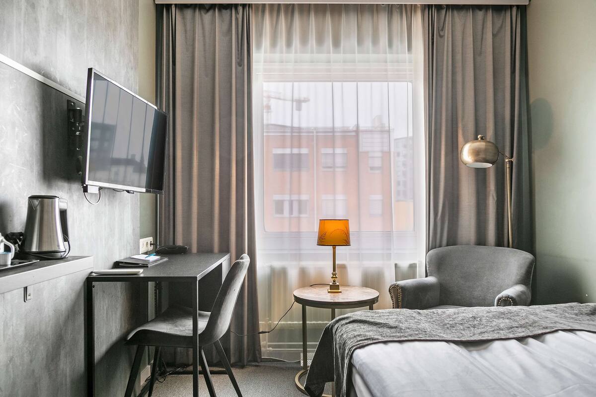 A hotel room with a bed, a desk with a TV, a chair, a lamp, and a view of buildings through the window.