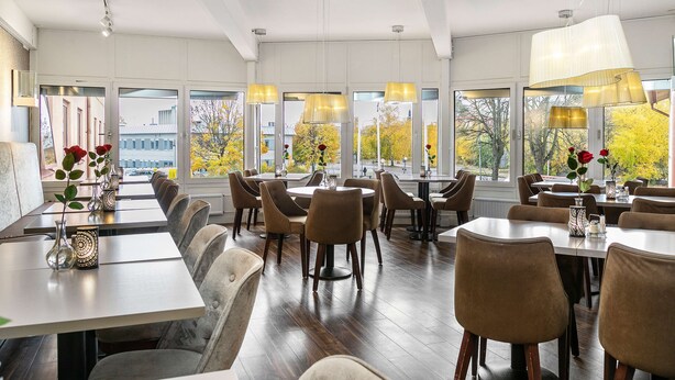 A spacious dining area with multiple tables and chairs, large windows with autumn foliage, and pendant lighting. - Hotel Mälarporten (Vasteras)
