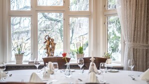 A dining table set with white tablecloth, plates, and wine glasses, with a view of trees and a statue through large windows. - Kronen Gaard Hotel (Sandnes)