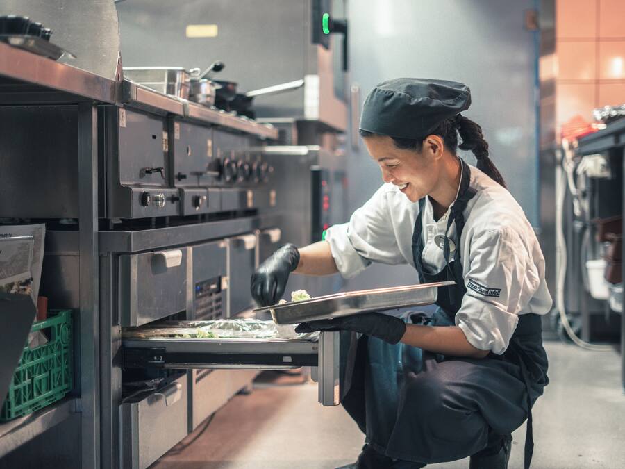 Een keuken met roestvrijstalen apparatuur, een kok met koksmuts en handschoenen, en een groene krat op het aanrecht.
