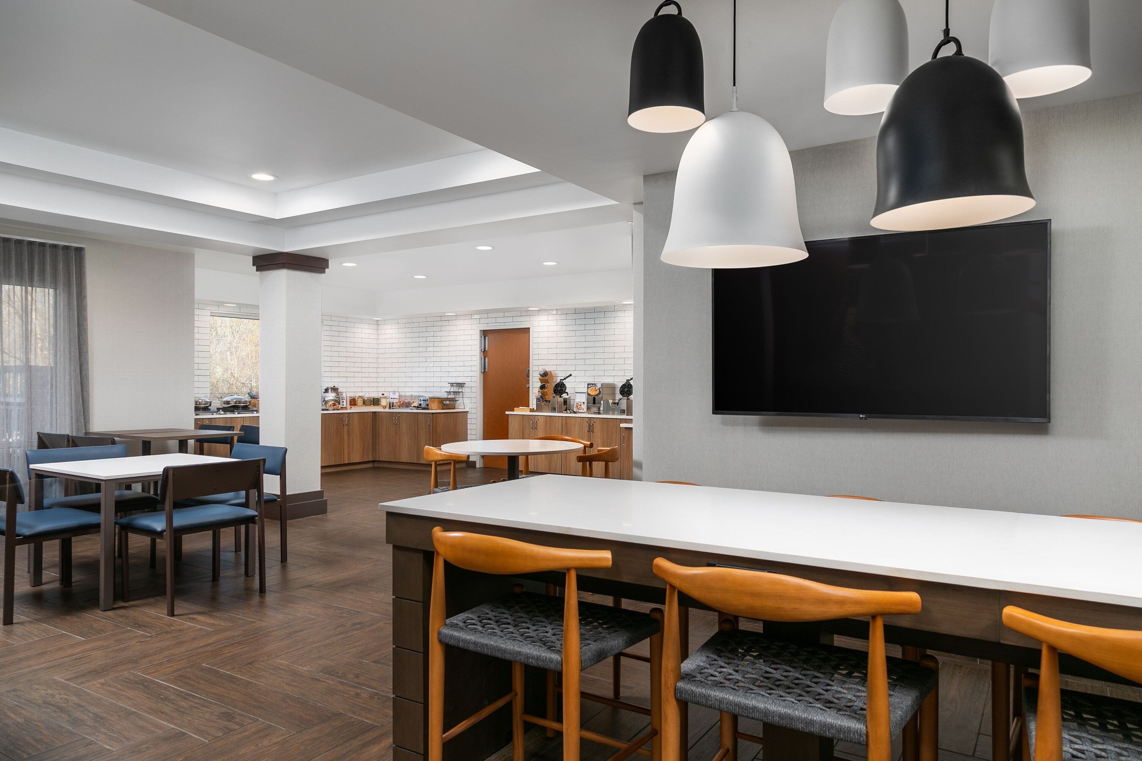Un espace repas moderne avec une longue table et des tabourets de bar, une cuisine en arrière-plan et un éclairage suspendu.