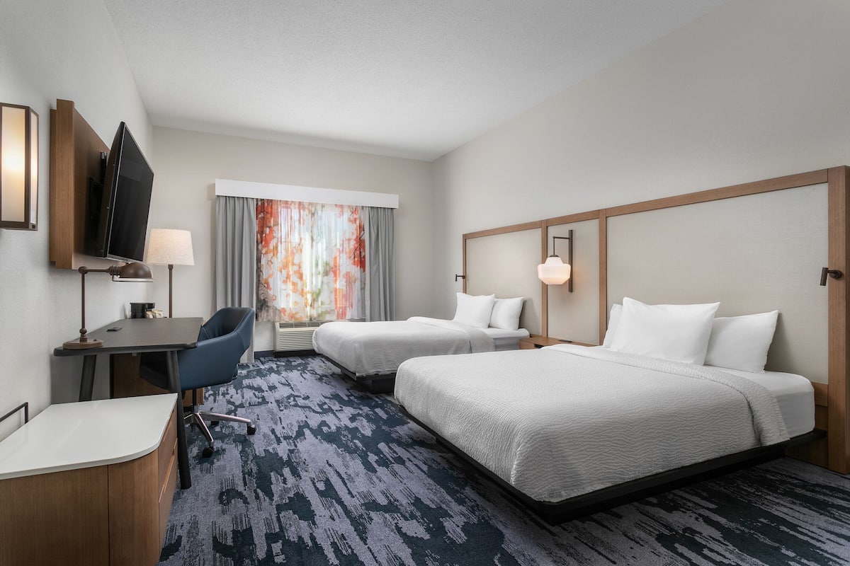A hotel room with two beds, a desk with a chair, a TV, and a window with curtains.