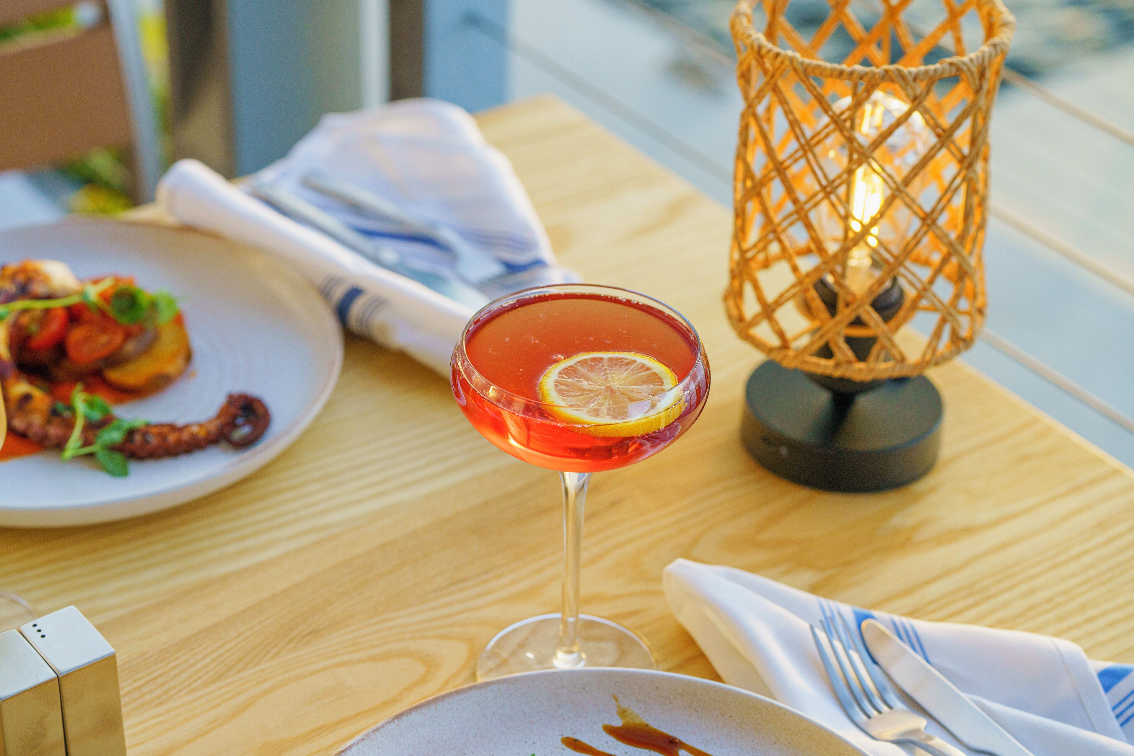Une table avec une assiette de nourriture, un verre à cocktail orné d’une tranche de citron et une lampe en osier.