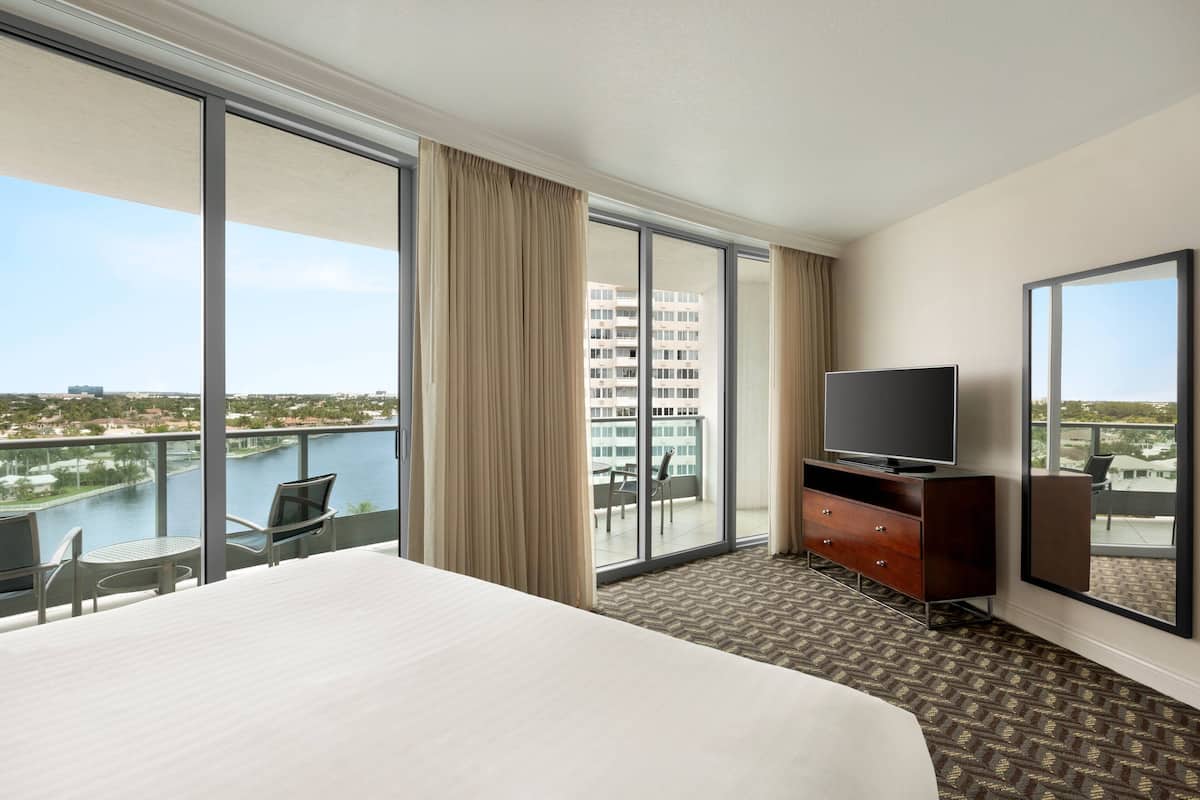 Habitación de hotel con una cama grande, un televisor sobre un aparador de madera y un balcón con vistas a un cuerpo de agua y edificios.