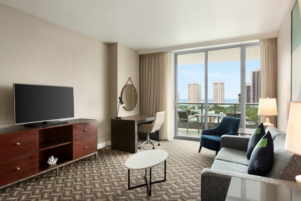Habitación de hotel con televisor de pantalla plana, escritorio con silla, sofá con cojines, mesa de centro y ventana grande con vista a la ciudad.