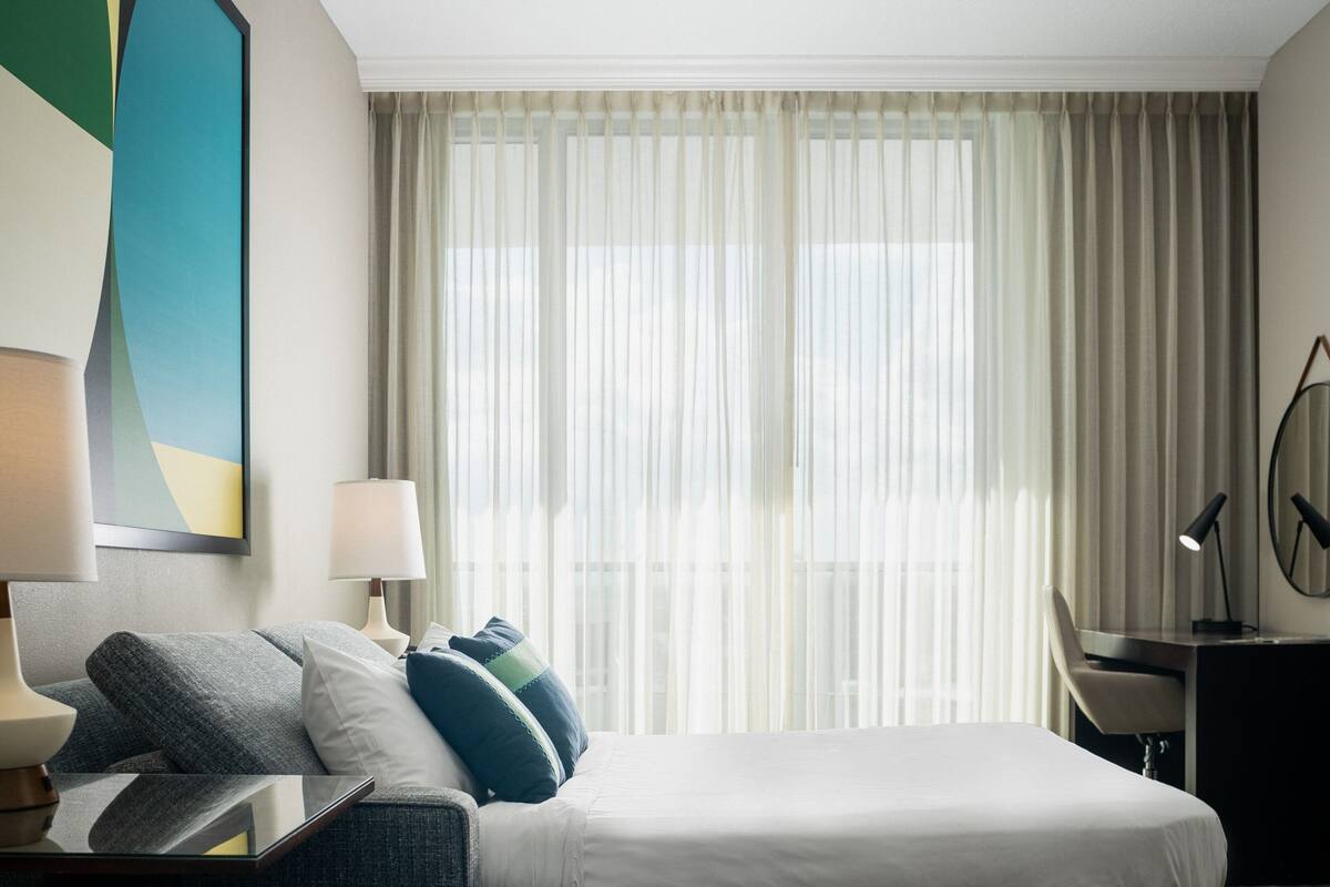 Habitación de hotel con cama, escritorio, silla, lámpara y una ventana grande con cortinas translúcidas.
