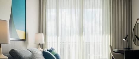 A hotel room with a bed, a desk, a chair, a lamp, and a large window with sheer curtains.