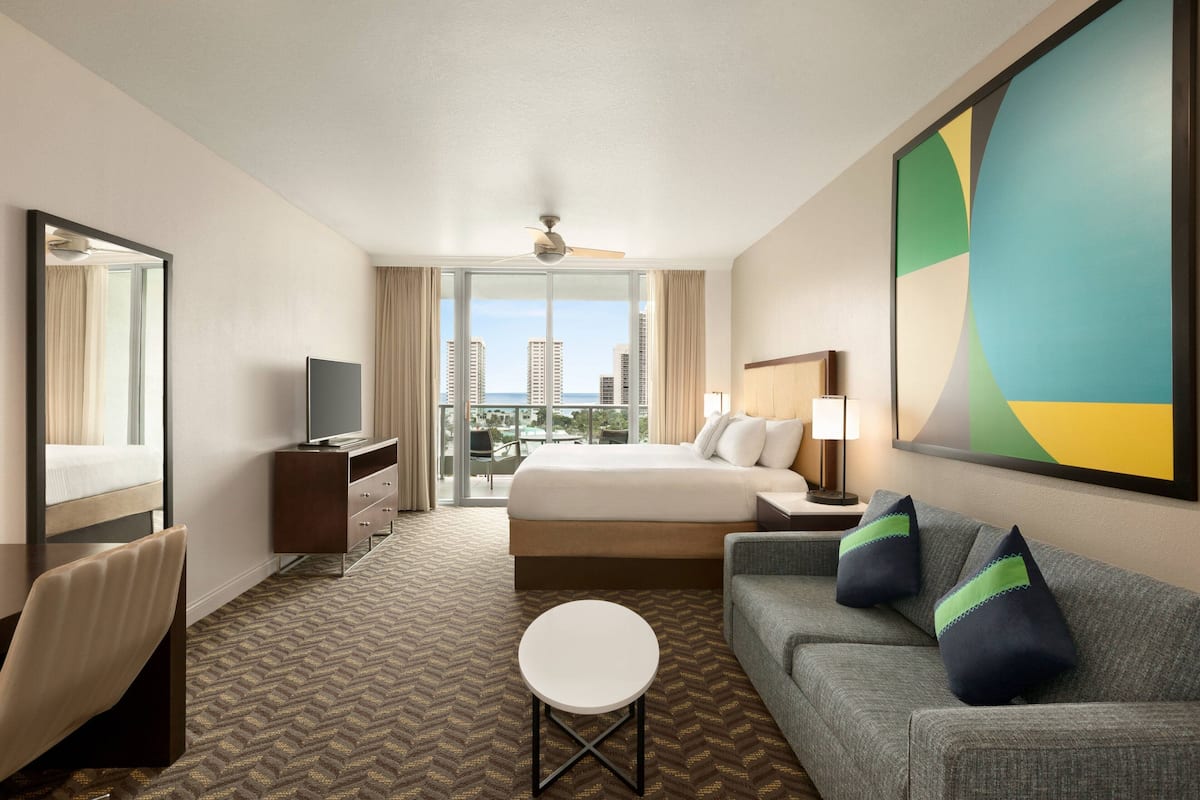 Habitación de hotel con cama, sofá, una mesita, televisión y un ventanal con vista a la ciudad.