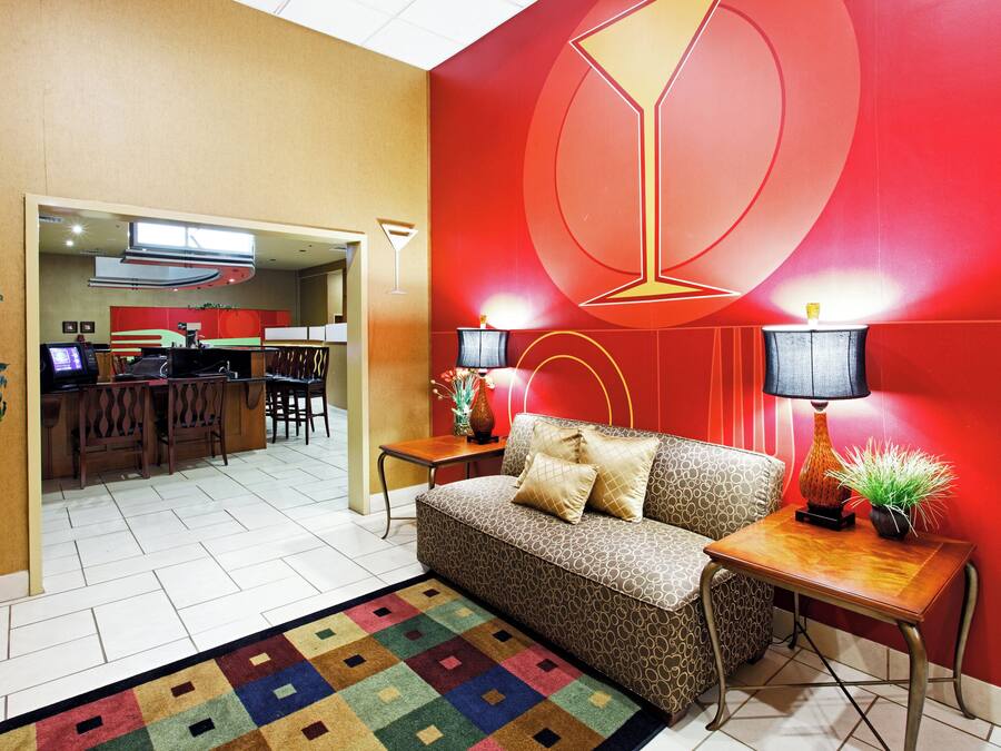 A modern interior with a red accent wall, a patterned sofa, a small table with a lamp, and a kitchen area in the background.