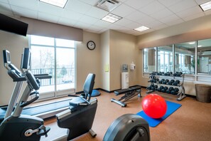 Een sportschool met loopbanden, elliptische trainers, een roeimachine, gewichten en een rode fitnessbal op een mat.