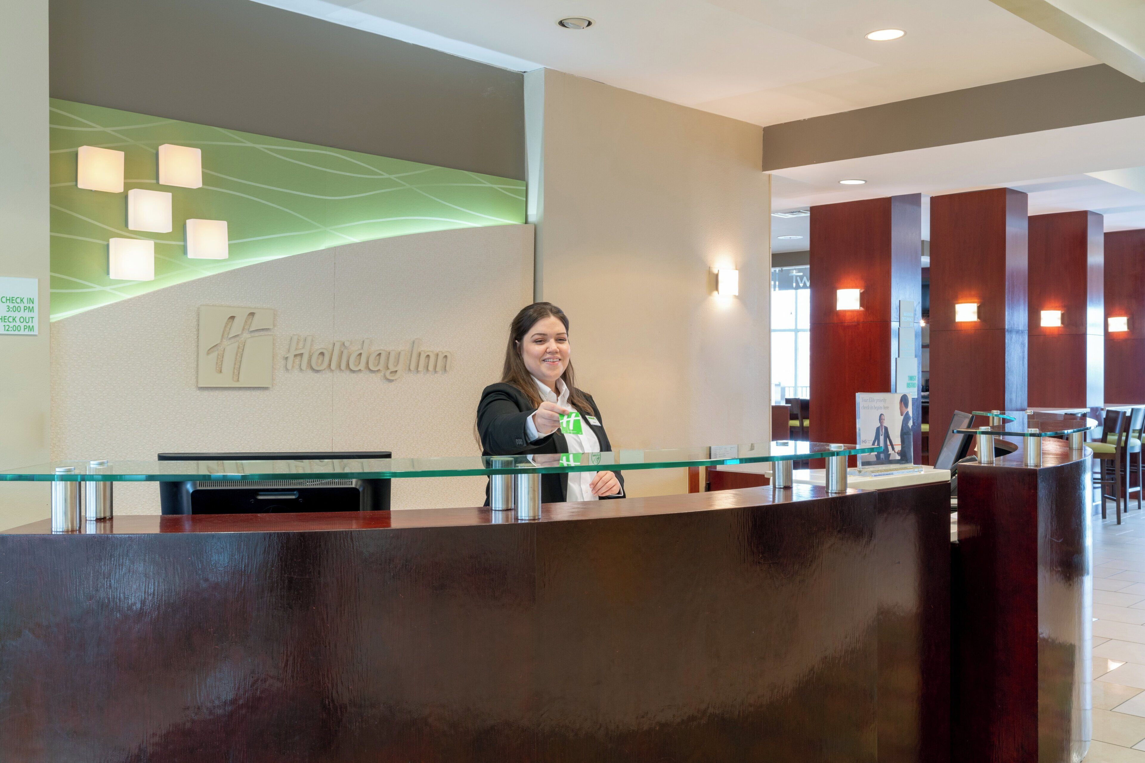 A reception desk at a Holiday Inn with a glass partition, a digital screen, and a sign indicating check-in and check-out times.