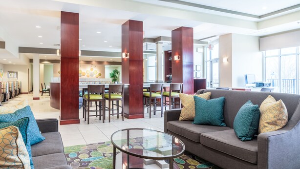 A spacious lobby with a gray sofa, a glass coffee table, and a patterned rug. There are wooden pillars and a dining area with chairs and a table in the background. - Holiday Inn Manassas - Battlefield by IHG (Manassas)