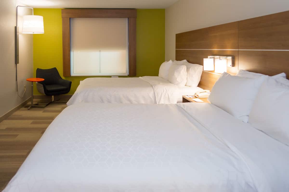 A hotel room with two beds, a chair, a lamp, and a window with blinds.