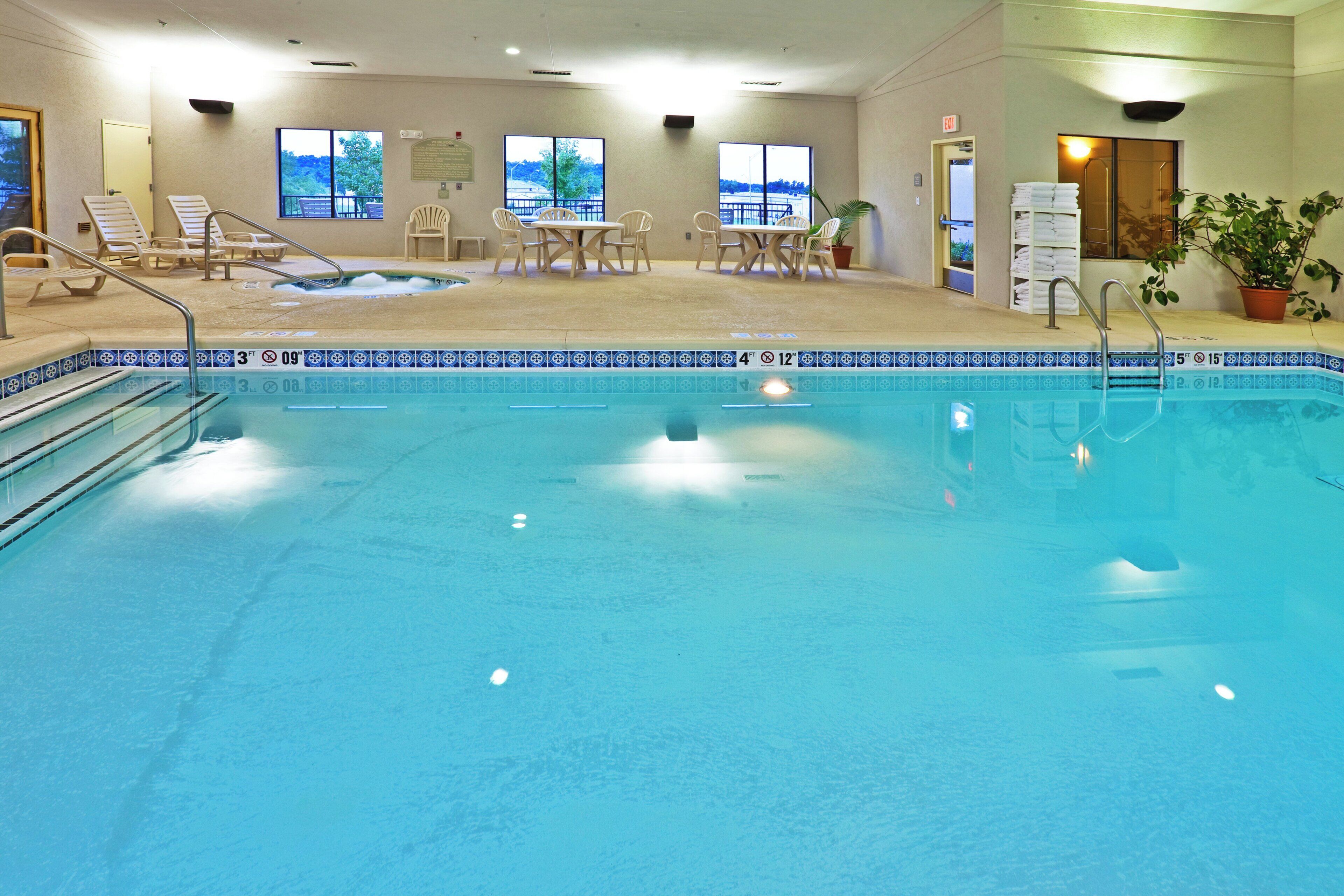An indoor swimming pool with a tiled edge, lounge chairs, a table with chairs, and a view of the outdoors through large windows.