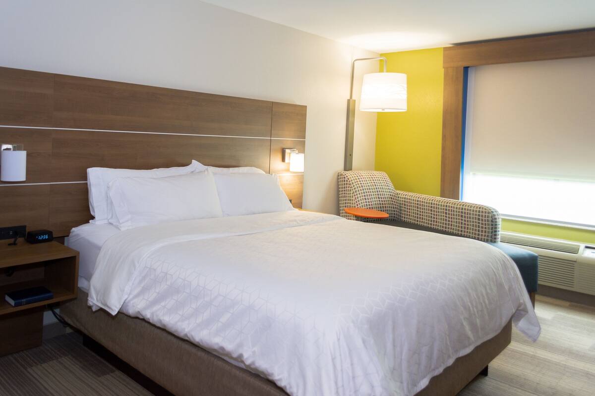 A hotel room with a large bed, a nightstand, a chair, and a window with blinds.