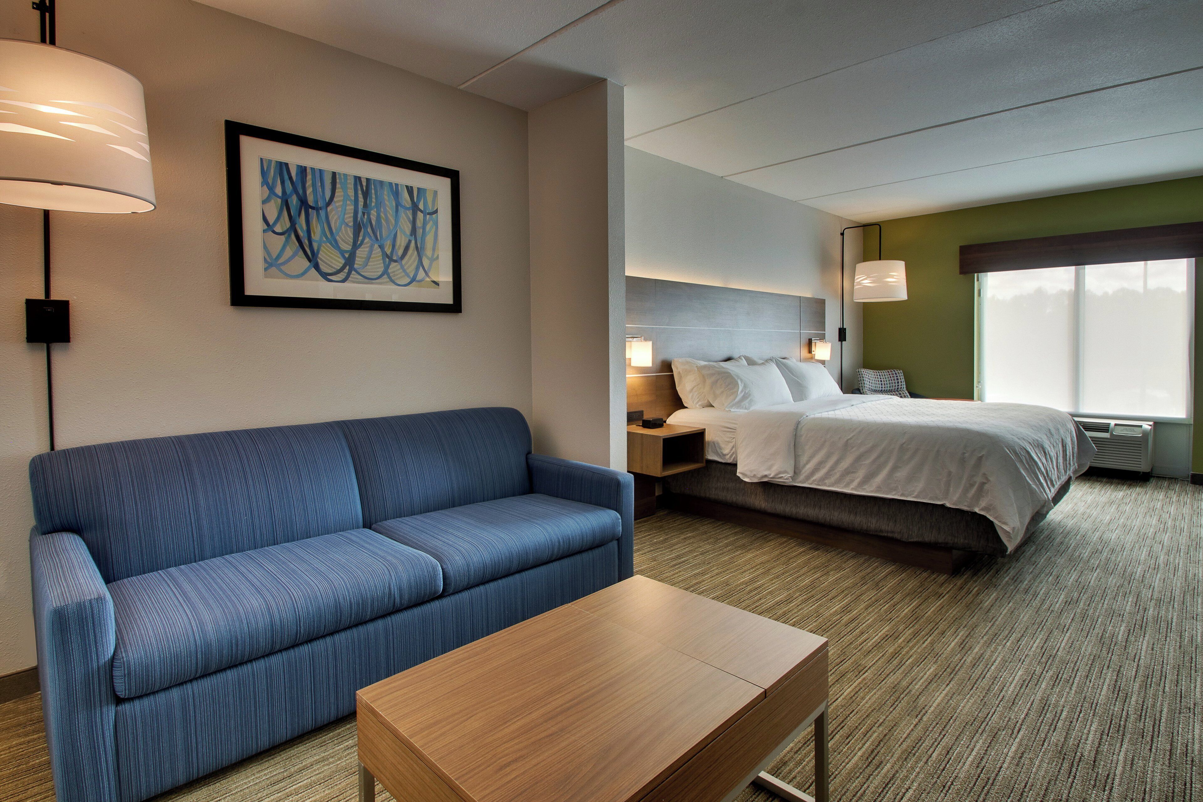 A hotel room with a blue sofa, a wooden table, a bed with white linens, a nightstand with a lamp, and a large window.