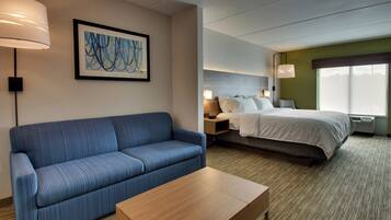 A hotel room with a blue sofa, a wooden table, a bed with white linens, a nightstand with a lamp, and a large window.