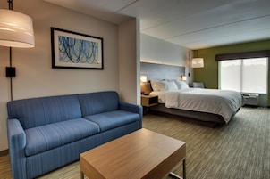 Habitación de hotel con un sofá azul, una mesa de madera, una cama con ropa blanca, una mesita de noche con una lámpara y un ventanal grande.