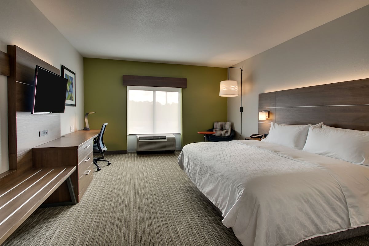 Ein Hotelzimmer mit Bett, Schreibtisch, Sessel, Wandfernseher und einem Fenster mit Jalousien.