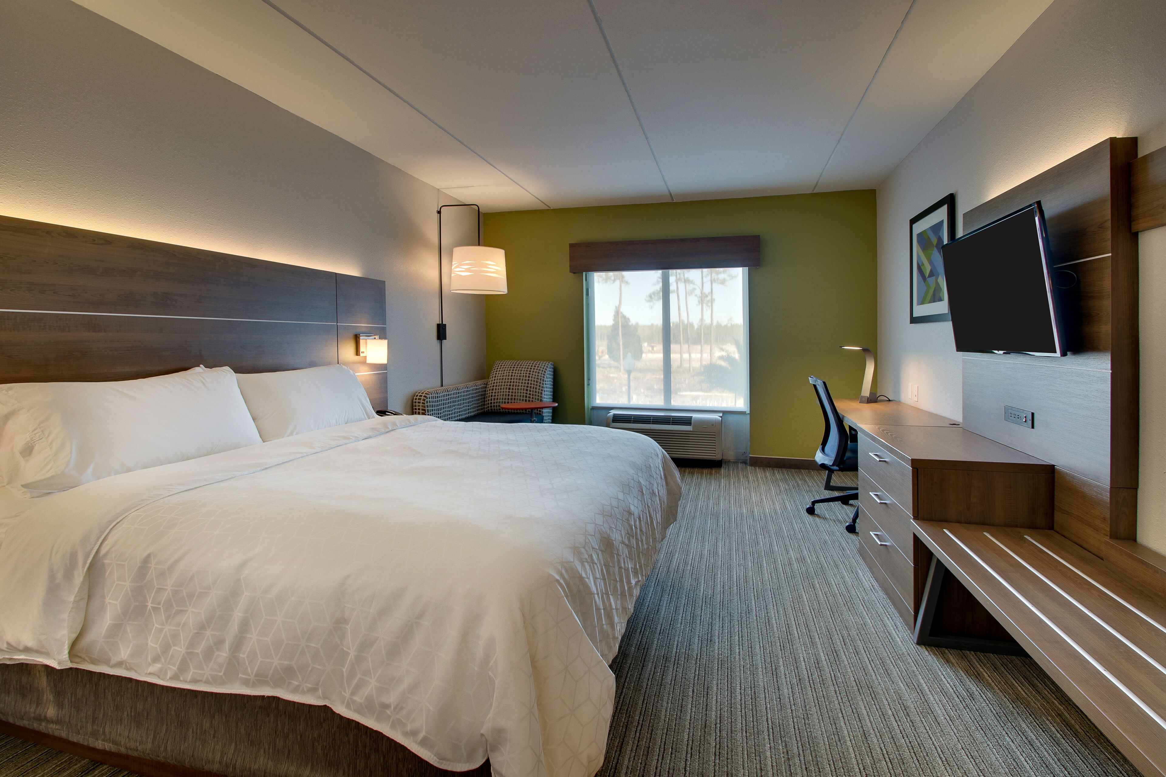 A hotel room with a large bed, a desk, a chair, a TV, and a window with a view of trees.