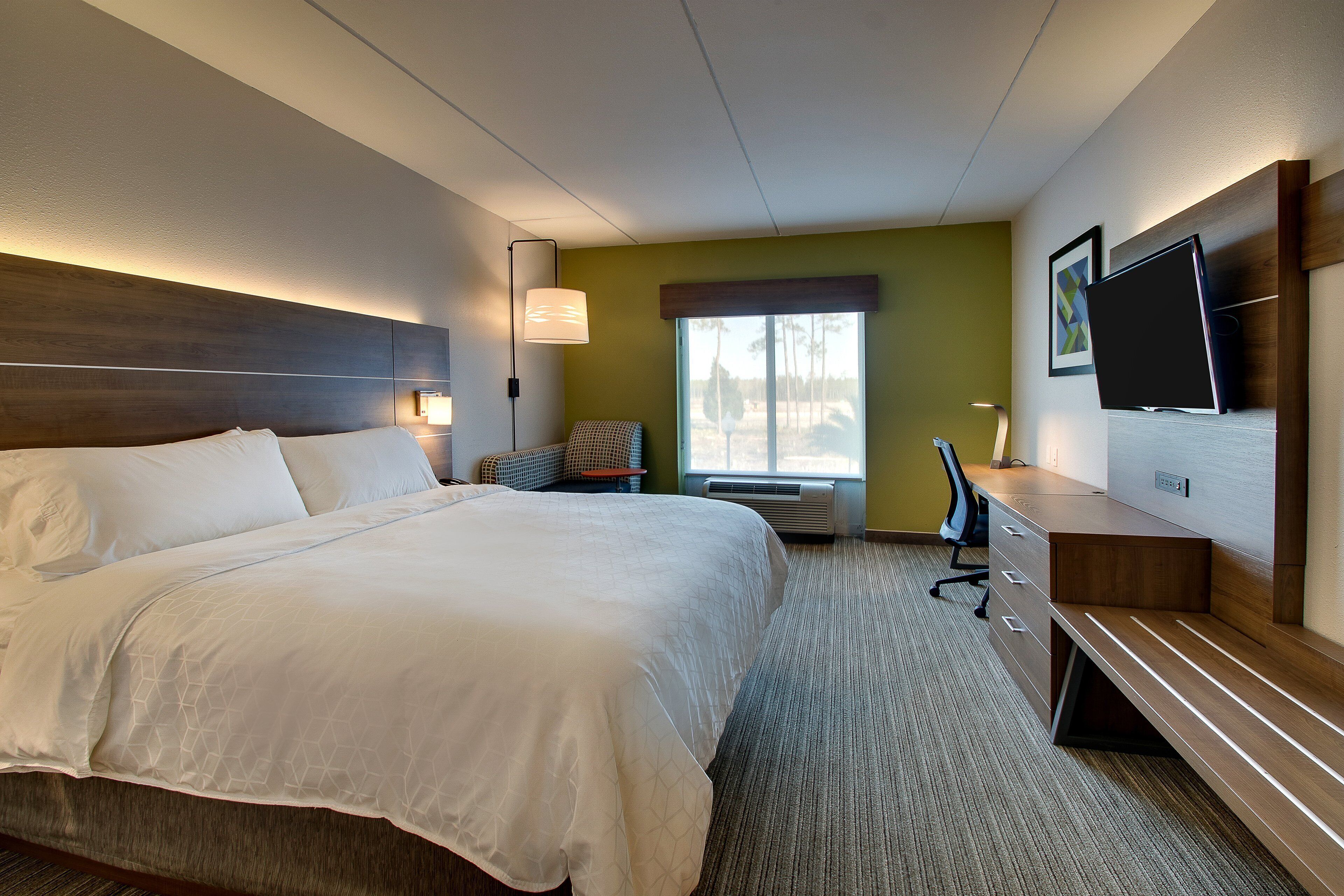 A hotel room with a large bed, a desk, a chair, a television, and a window with a view of trees.