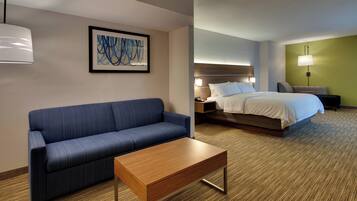 A hotel room with a blue sofa, a wooden coffee table, a bed with white bedding, a nightstand with a lamp, and a framed abstract painting on the wall.