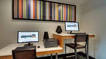 A hotel room with two desks, each with a computer monitor, keyboard, and mouse. There is a printer on the desk, and a chair is positioned in front of the desk. A large, colorful striped artwork is displayed on the wall above the desks.