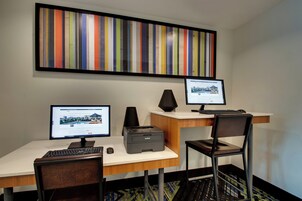 Habitación de hotel con dos escritorios, cada uno con un monitor de computadora, teclado y ratón. Hay una impresora en el escritorio, y una silla está colocada frente a él. Una llamativa obra de arte a rayas está expuesta en la pared sobre los escritorios.