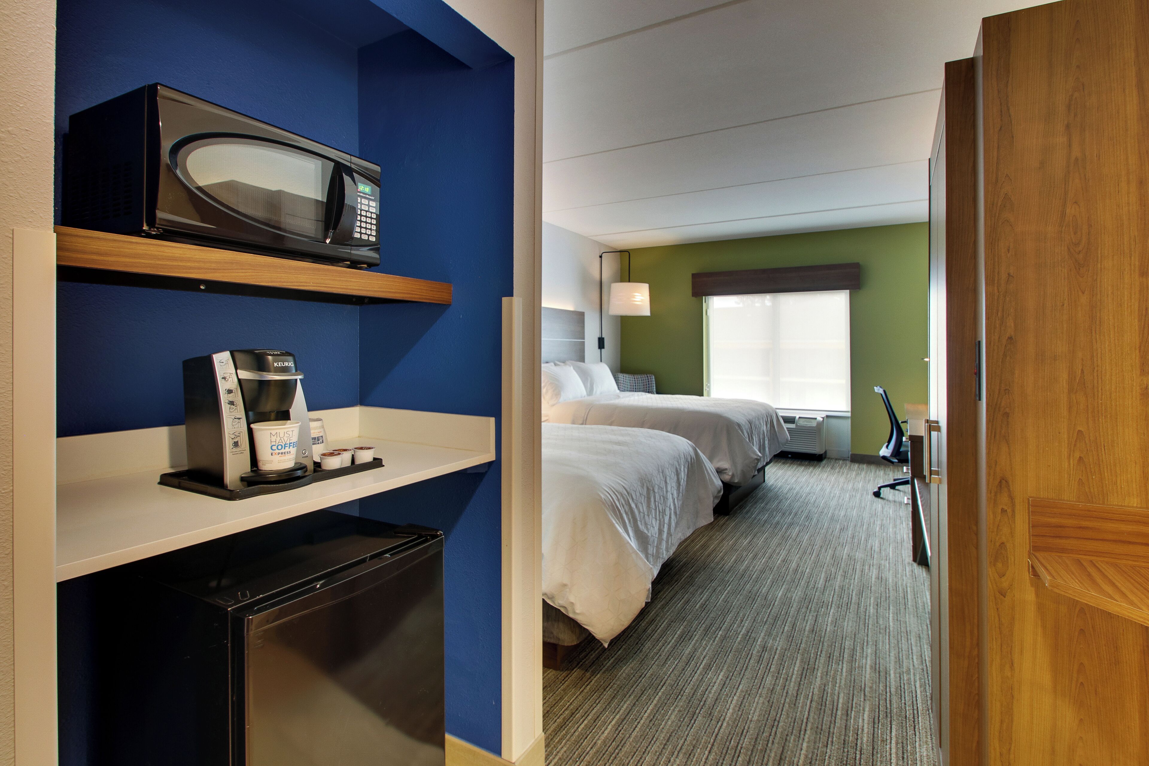Habitación de hotel con kitchenette, una cama, una mesita de noche, una lámpara y una ventana.