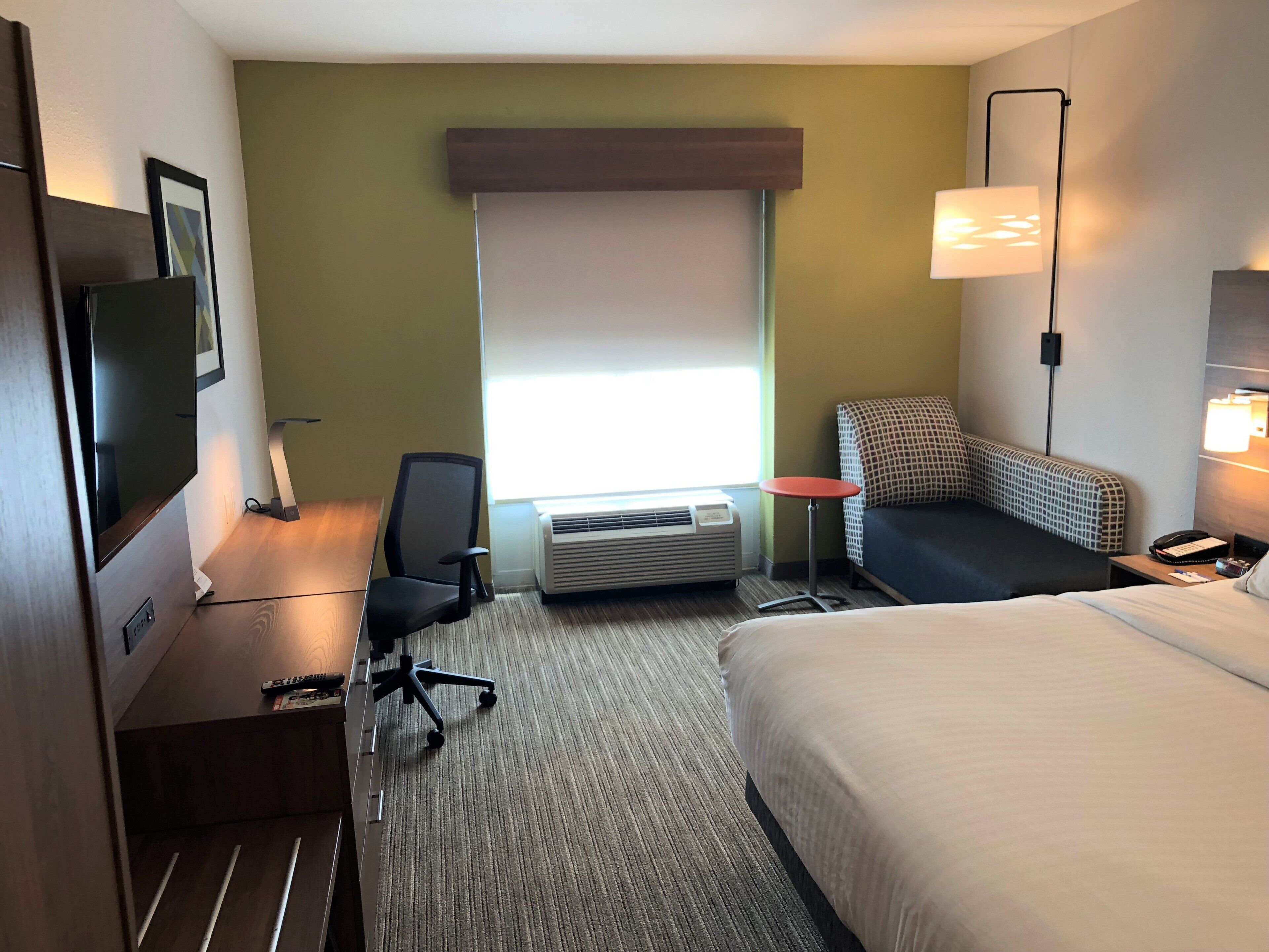 Ein Hotelzimmer mit Bett, Schreibtisch mit Stuhl, einem kleinen Tisch, einem Wand-Fernseher, einem Fenster mit Jalousien und einer Stehlampe.