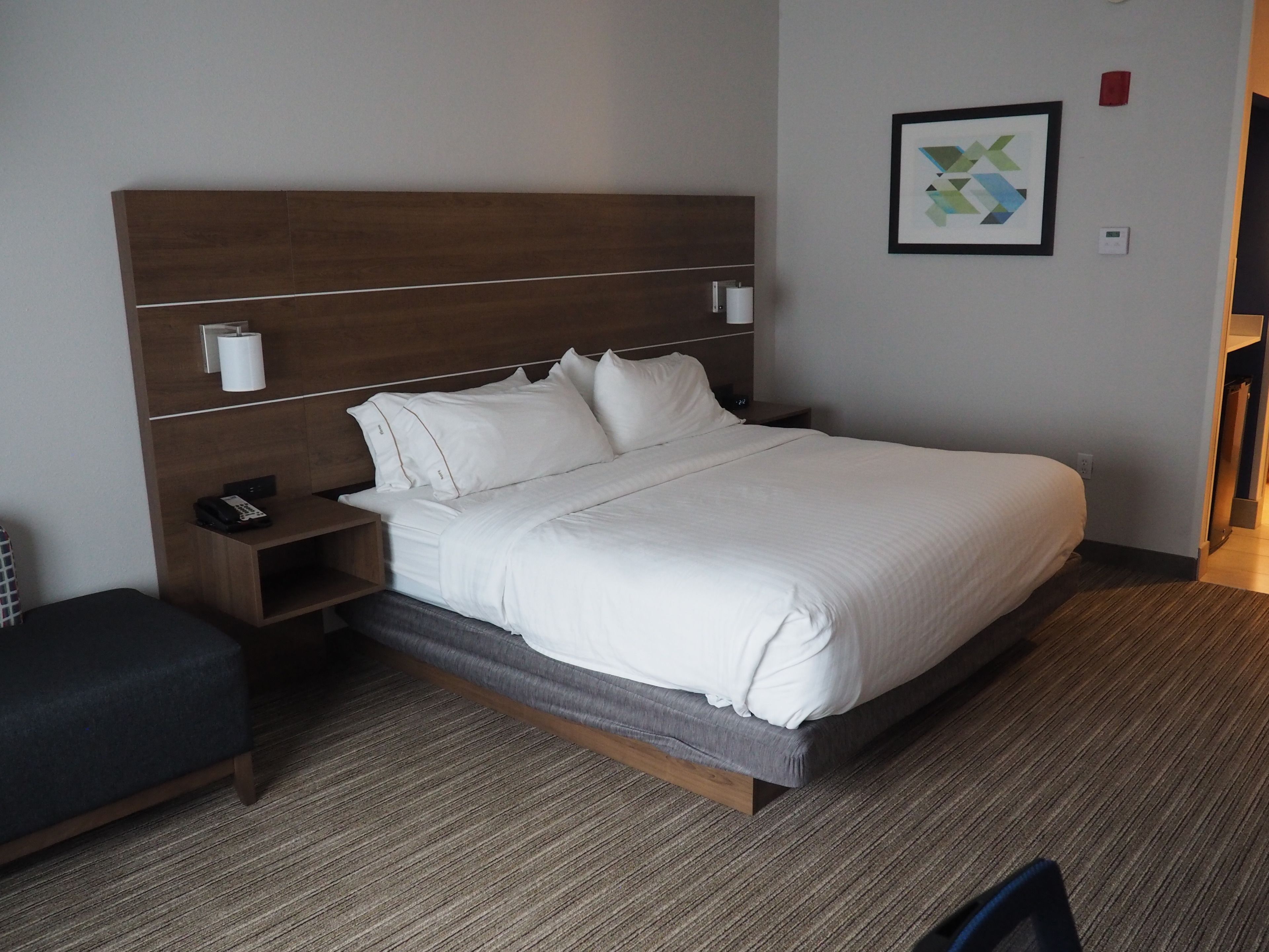 Una habitación de hotel con una cama grande, una mesita de noche con un teléfono, una lámpara de pared y un cuadro en la pared.