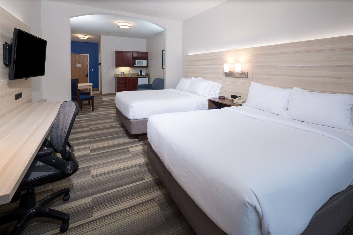 A hotel room with two beds, a desk, a chair, a TV, a microwave, and a kitchenette.