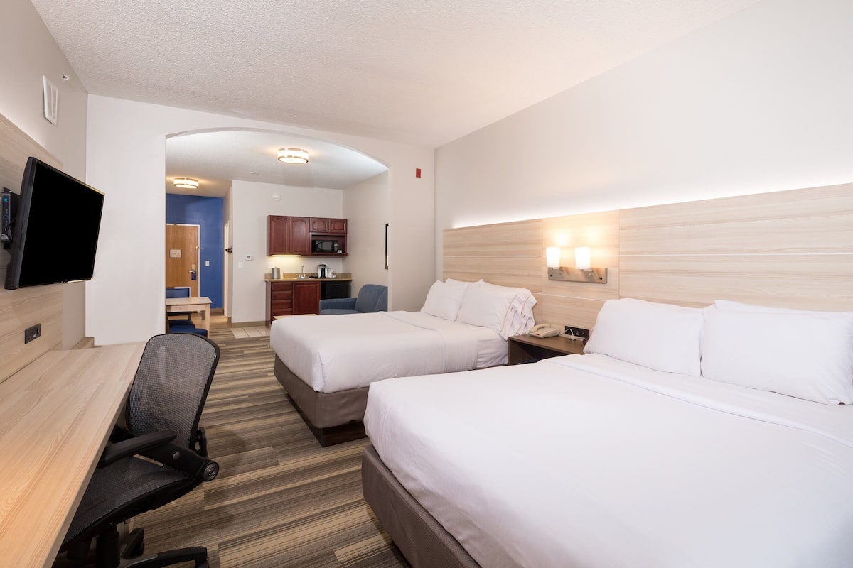 A hotel room with two beds, a desk, a TV, a microwave, and a small kitchen area.