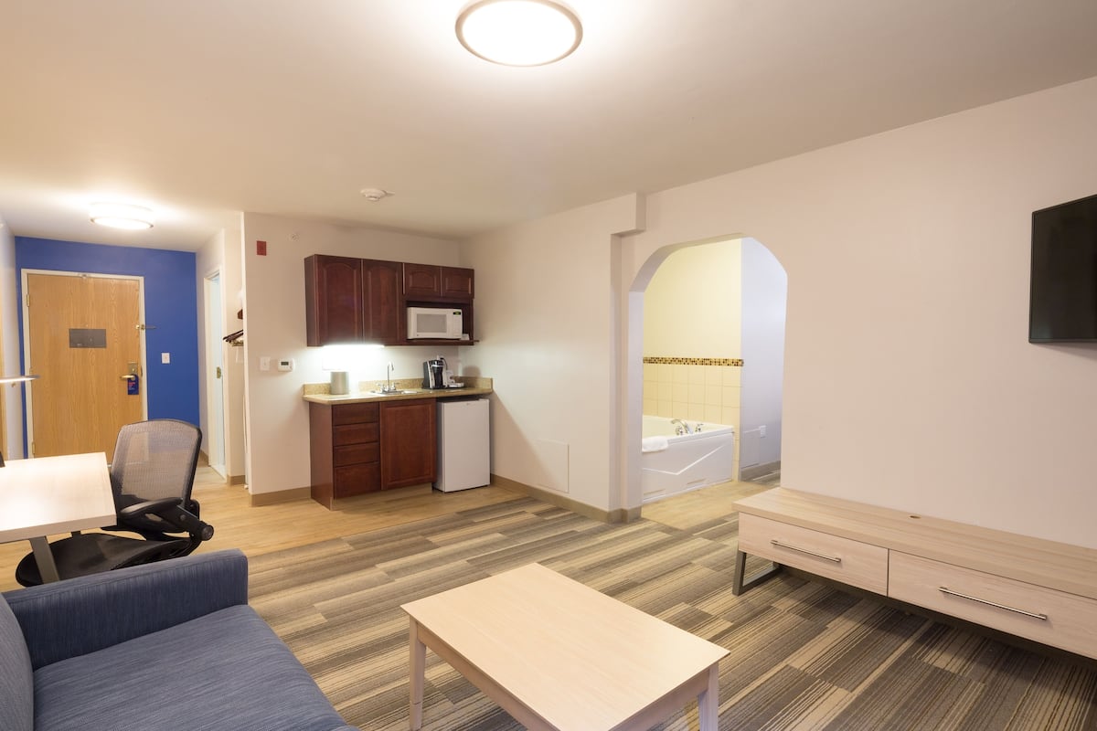 A modern hotel room with a kitchenette, a seating area with a table and chairs, and a large mirror.