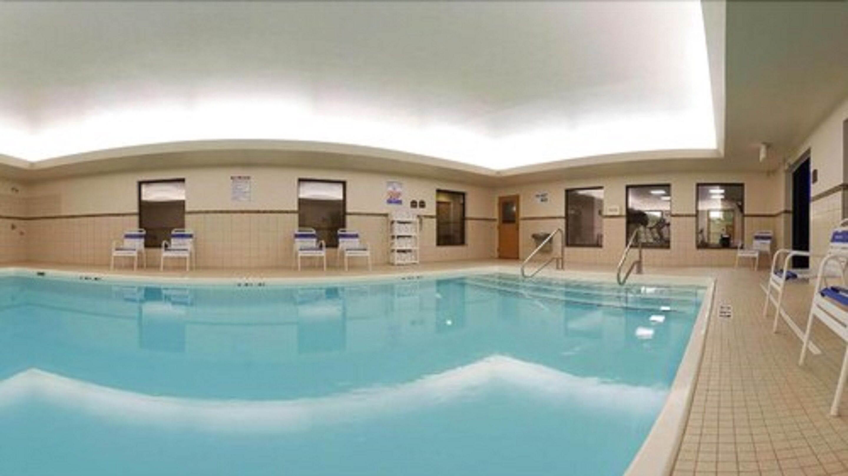 An indoor swimming pool with clear water, surrounded by tiled walls and a tiled floor. There are several lounge chairs and a pool ladder visible.