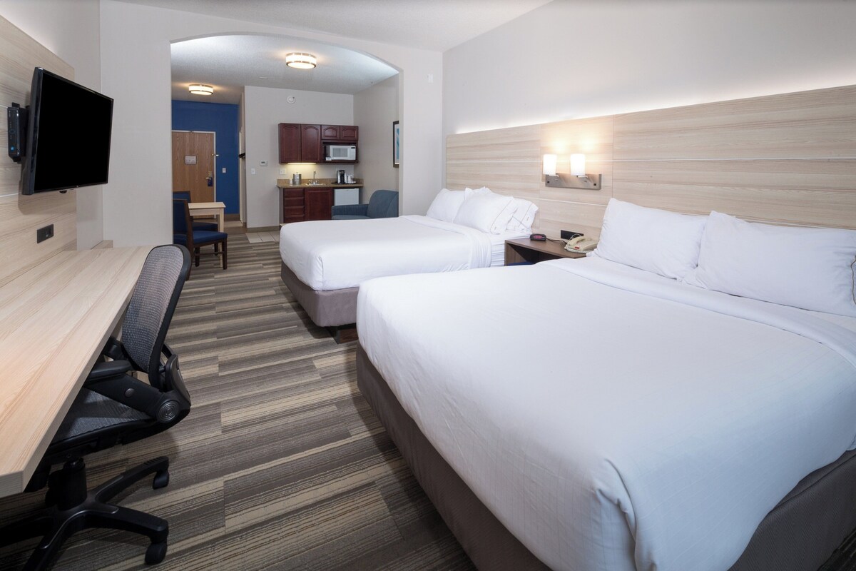 A hotel room with two beds, a desk, a chair, a TV, a microwave, and a kitchenette.
