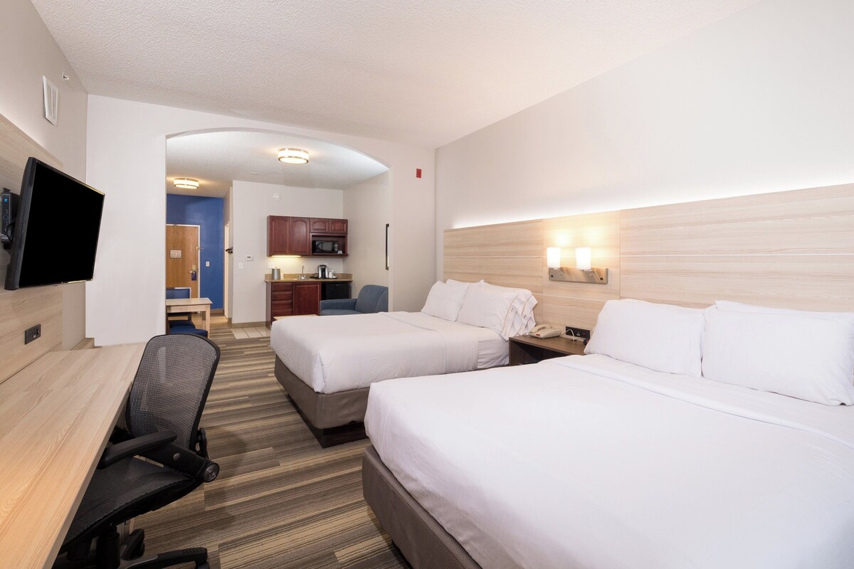 A hotel room with two beds, a desk, a chair, a TV, and a kitchenette.