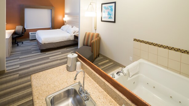 A hotel room with a bed, a desk, a chair, a bathroom with a sink and bathtub, and a mirror. - Holiday Inn Express Hotel & Suites Grand Blanc by IHG (Grand Blanc)