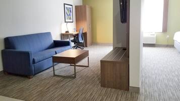 A hotel room with a blue sofa, a wooden coffee table, a desk with a chair, a wardrobe, and a bed.
