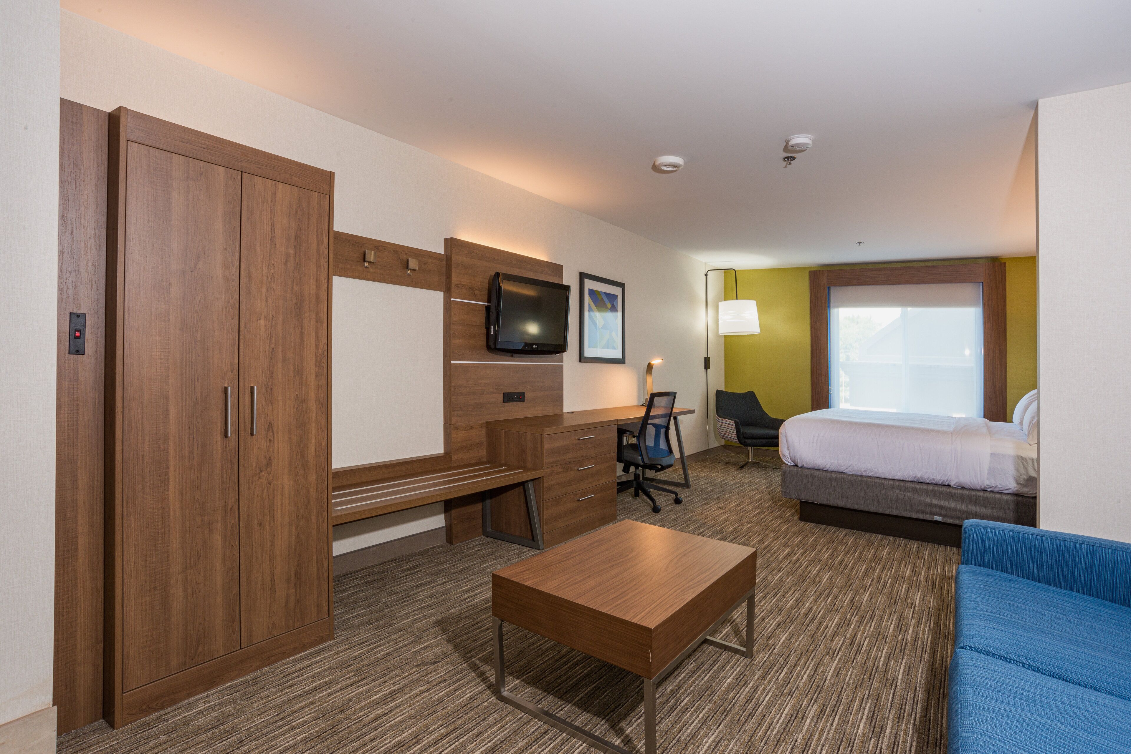 A hotel room with a wooden wardrobe, a desk with a chair, a bed, a blue sofa, and a television on the wall.