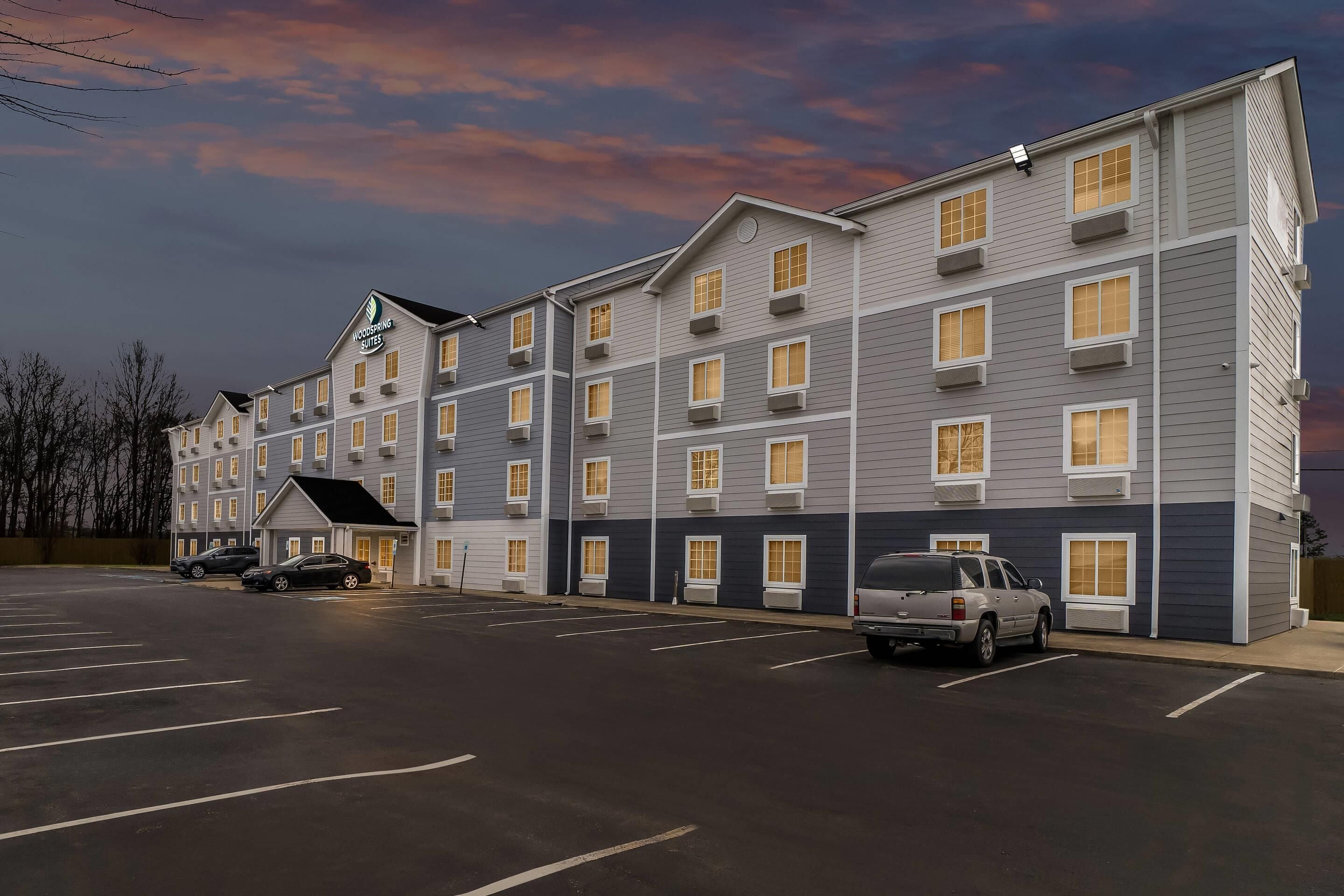 Un hôtel à plusieurs étages avec un parking, plusieurs voitures garées et un ciel dégagé.