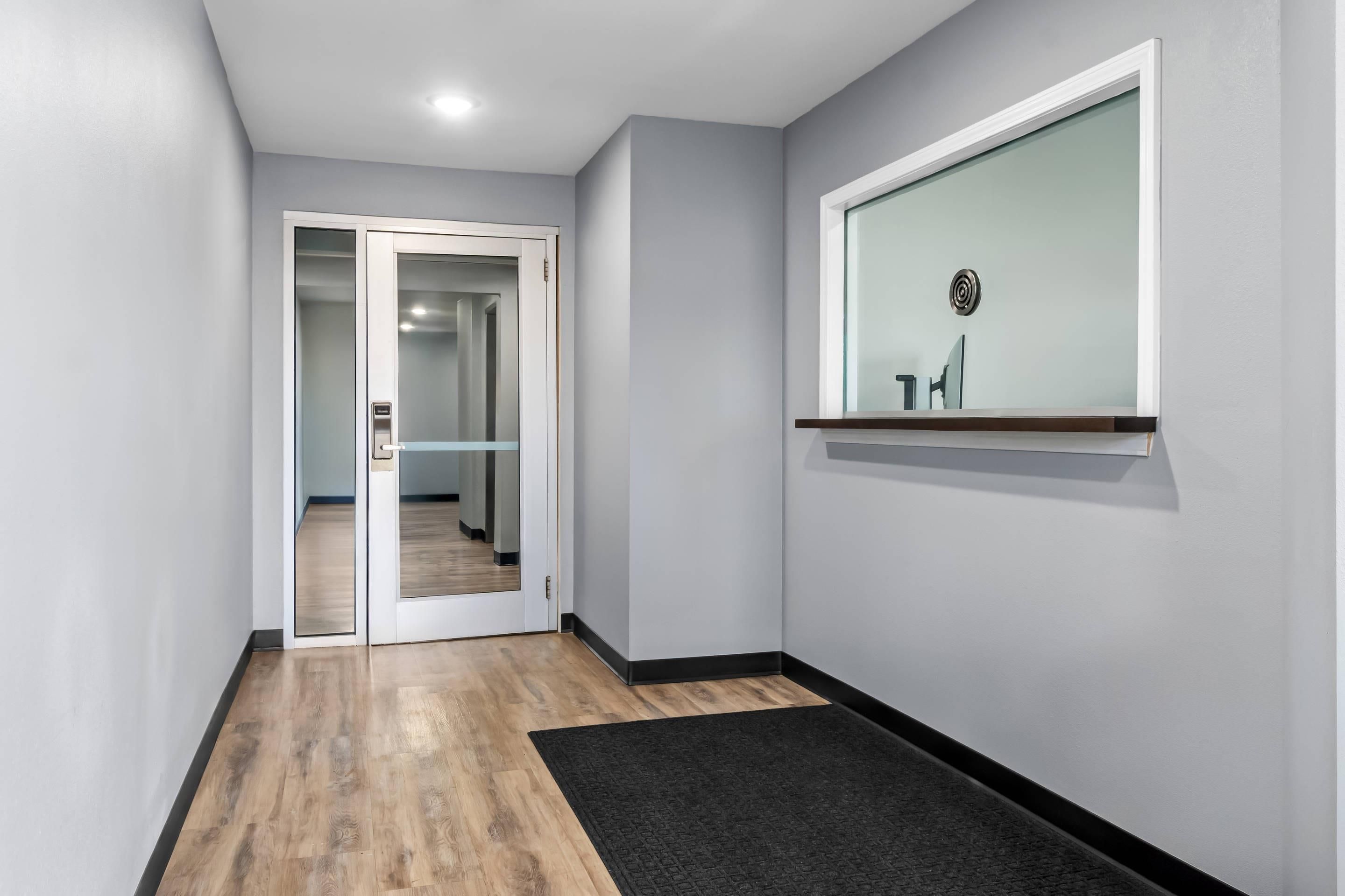 A hallway with a wooden floor, a black doormat, a glass door leading to another room, and a wall-mounted shelf with a small item.