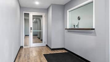A hallway with a wooden floor, a black doormat, a glass door leading to another room, and a wall-mounted shelf with a small item.