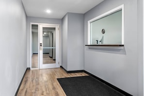 A hallway with a wooden floor, a black doormat, a glass door leading to another room, and a wall-mounted shelf with a small item. - WoodSpring Suites Memphis East I-40 (Memphis)