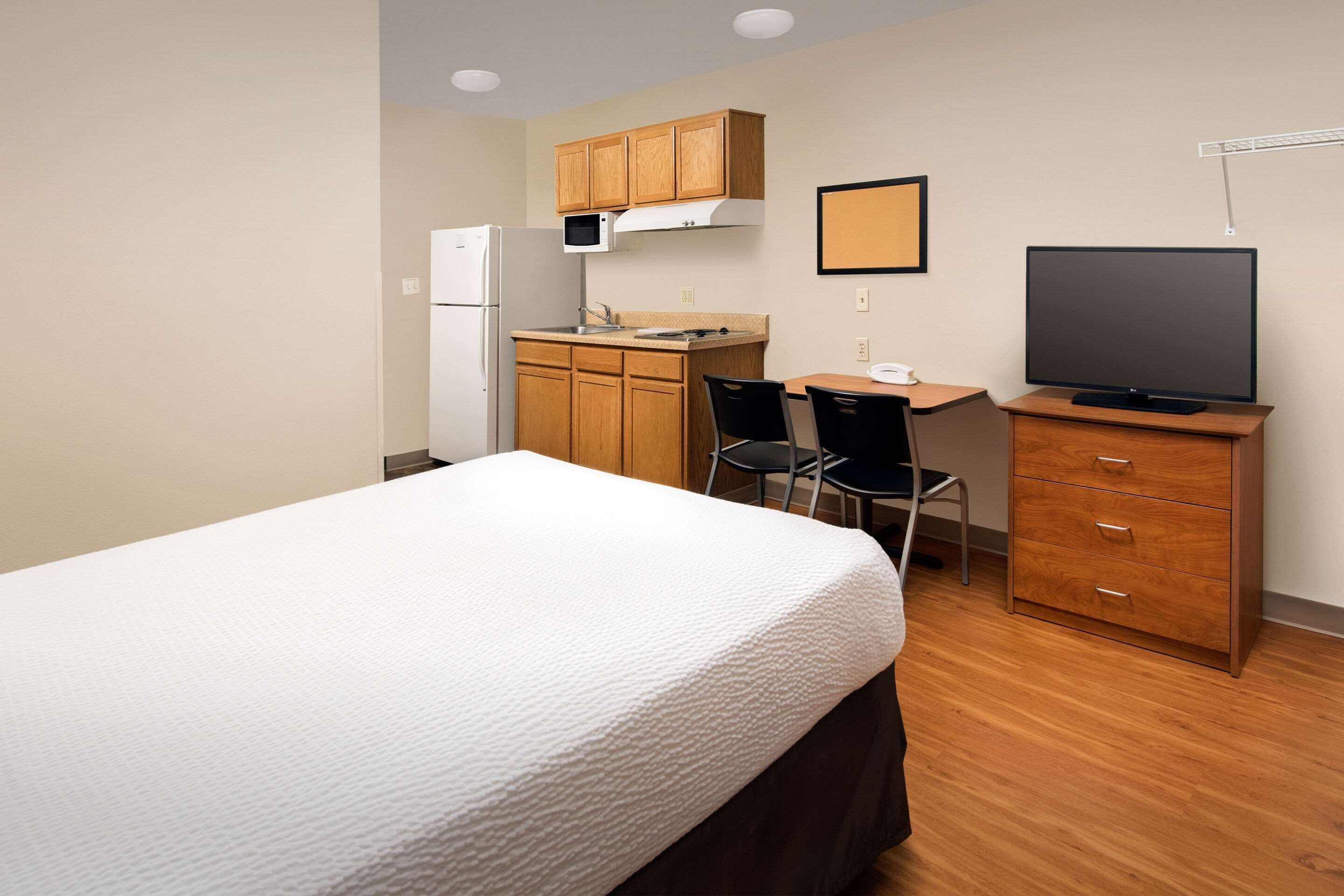 A compact room with a bed, a kitchenette, a desk with a chair, and a television.