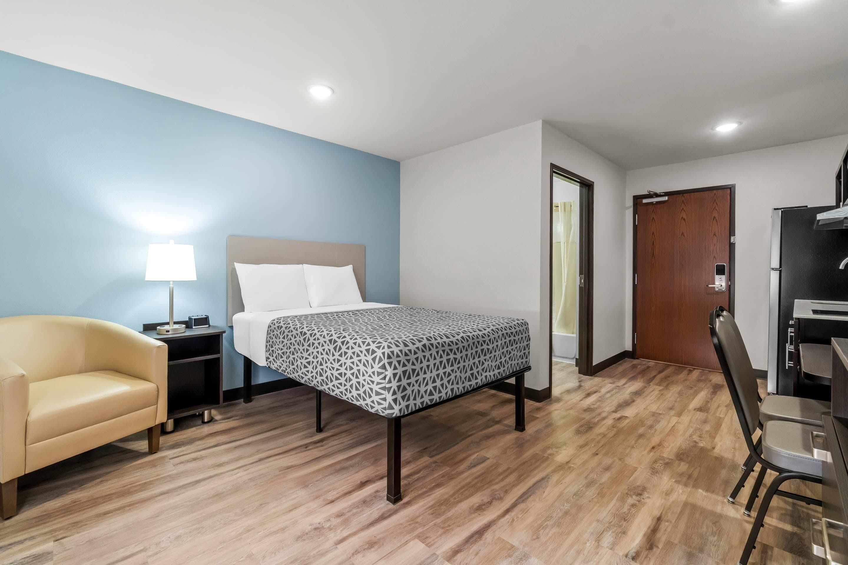 A hotel room with a bed, a chair, a desk, a lamp, and a door.