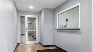 Ein Flur mit Holzboden, einer schwarzen Fußmatte, einer Glastür zu einem anderen Raum und einem an der Wand befestigten Regal mit einem kleinen Gegenstand.