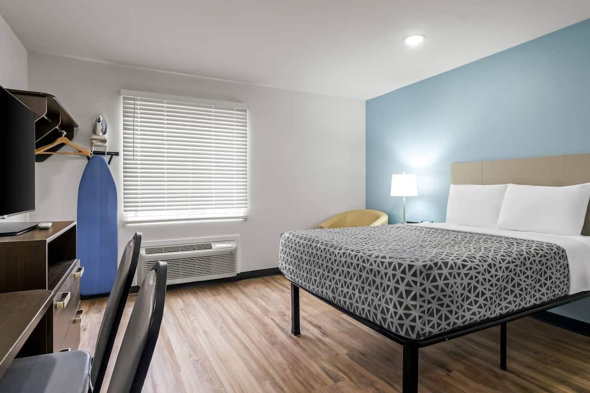 A hotel room with a bed, desk, chair, and a window with blinds.